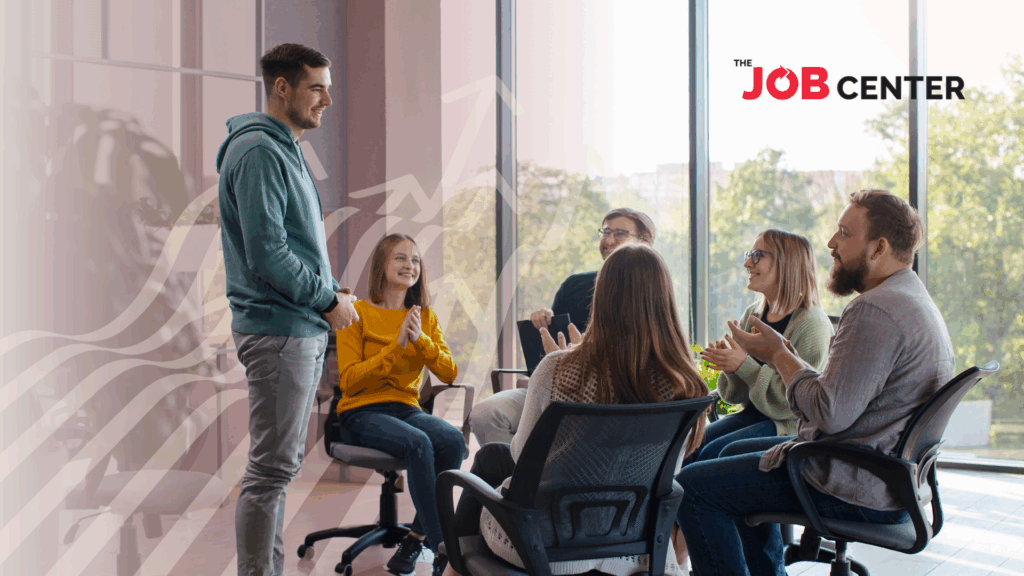 A group of employees sits in a circle of office chairs, applauding one standing employee